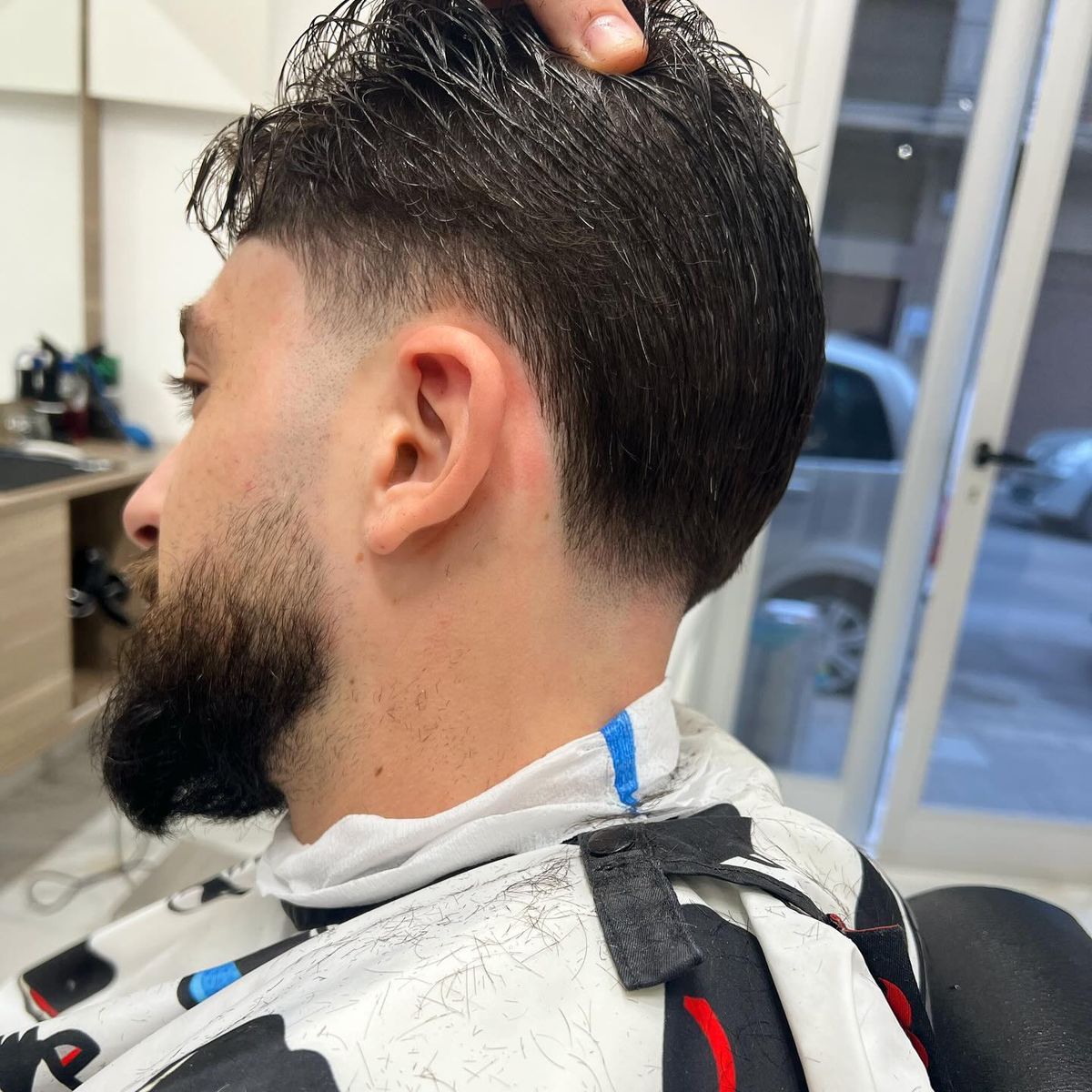 Low fade e barba piena, capelli pettinati all’indietro, di profilo